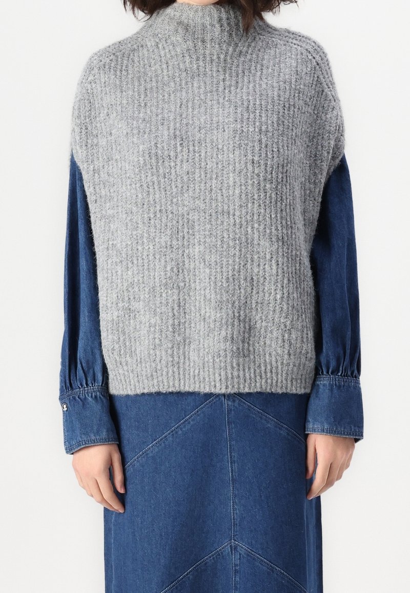 Personne portant un pull sans manches en tricot gris par-dessus une robe en jean bleue à manches longues avec des poignets boutonnés, debout devant un fond blanc.