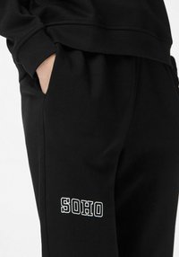 Czarne spodnie dresowe o luźnym kroju, z boczną kieszenią i napisem "SOHO" w białym kolorze na dolnej części nogawki. Miękki materiał bawełniany.