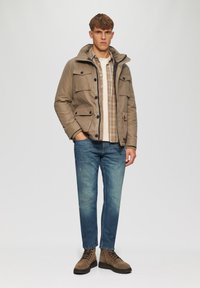 Khaki hoodie met knopen- en zakdetails, gedragen over een geruit overhemd. Gecombineerd met lichtblauwe jeans en bruine veterschoenen.
