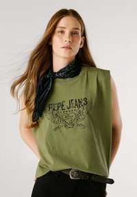 Canottiera senza maniche verde oliva con un logo stampato nero che presenta delle ali, indossata con una sciarpa nera e una cintura nera, che mostra un fit rilassato.
