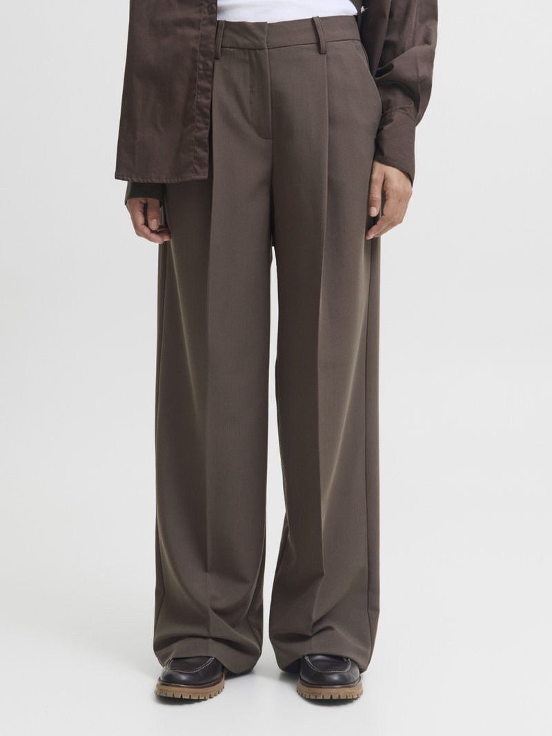 Calças largas castanhas com uma textura de tecido suave, apresentando uma cintura ajustada, pregas e um design limpo e bem cortado. Combinadas com sapatos escuros.