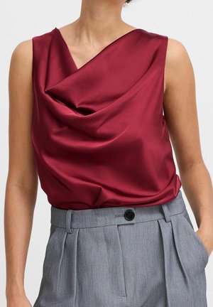Czerwona satynowa bluzka bez rękawów z drapowaniem, wsunięta w wysokowarstwowe, szare, dopasowane spodnie z czarnym guzikiem i szlufkami na pasek.