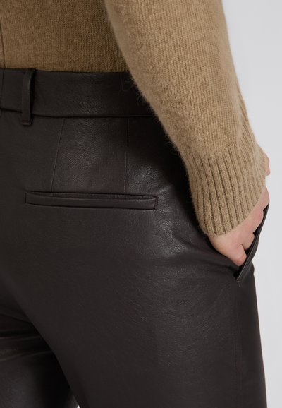 Brązowe skórzane spodnie o gładkiej fakturze, z boczną kieszenią i dopasowanym paskiem w talii, zestawione z beżowym swetrem z dzianiny.