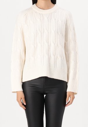 Femme portant un pull en maille torsadée blanc et un pantalon en cuir noir, debout devant un fond blanc uni.