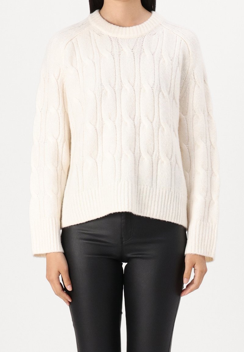 Femme portant un pull en maille torsadée blanc et un pantalon en cuir noir, debout devant un fond blanc uni.