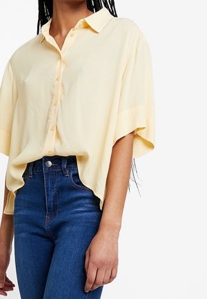 Camisa amarilla de manga corta con botones, hecha de tela ligera, con un corte holgado y cuello, combinada con vaqueros azules de talle alto.