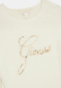Pull duveteux couleur crème avec "Guess" écrit sur le devant en sequins dorés brillants et une petite étiquette de marque près de l'encolure.