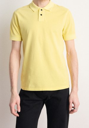 Homme portant un polo à manches courtes jaune clair avec deux boutons noirs et un pantalon noir, debout devant un fond neutre.