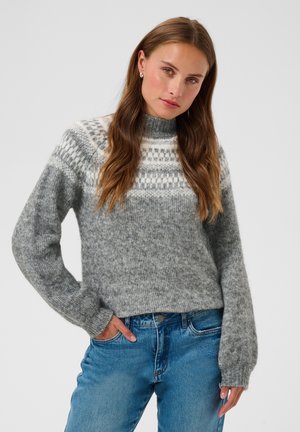 Jeune femme aux longs cheveux bruns portant un pull en maille gris à motifs et un jean bleu, debout avec une main dans sa poche.