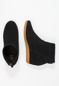 Bottines en daim noir avec texture perforée, semelle en caoutchouc et fermeture éclair latérale. Elles présentent un embout arrondi et une hauteur à la cheville.