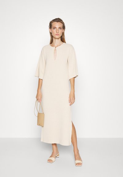 Μπεζ πλεκτό midi φόρεμα με λαιμόκοψη σε σχήμα κλειδώματος, κοντά φουσκωτά μανίκια, πλαϊνή σχισμή και απαλό υφή· συνδυασμένο με μπεζ σαγιονάρες και τσάντα.
