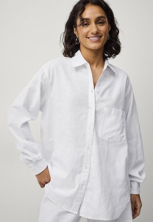 Chemise en lin blanche à manches longues, avec un bouton sur le devant, un col pointu et une poche poitrine unique. Présente une coupe décontractée et une texture légère.