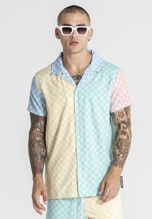 Mand iført pastelternet kortærmet skjorte med knapper og matchende shorts, hvide solbriller, med tatoveringer på begge arme og bryst.