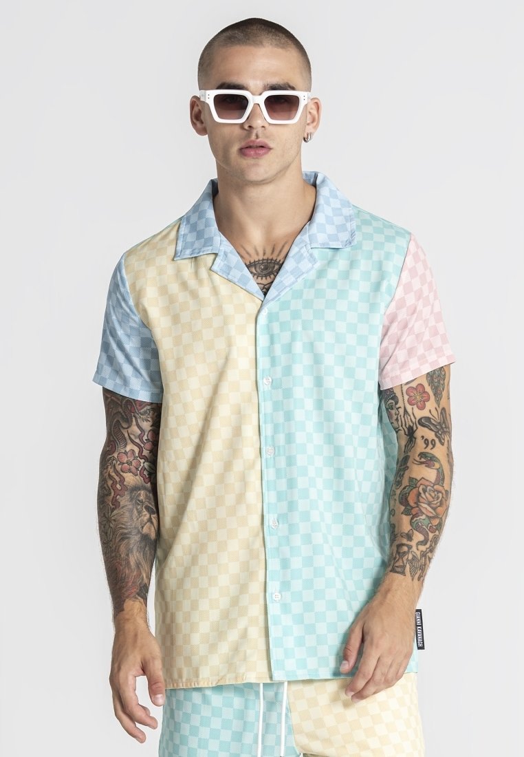 Uomo con camicia a maniche corte abbottonata a quadri pastello e pantaloncini abbinati, occhiali da sole bianchi, con tatuaggi su entrambe le braccia e il petto.