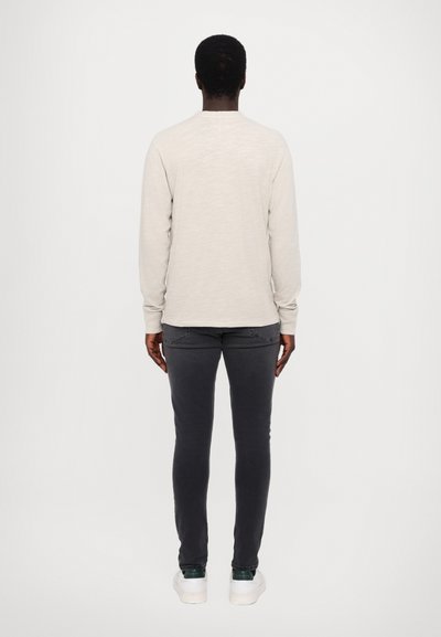 Lys beige langermet skjorte med rund hals, kombinert med mørkegrå slim-fit jeans og hvite joggesko med grønne detaljer.