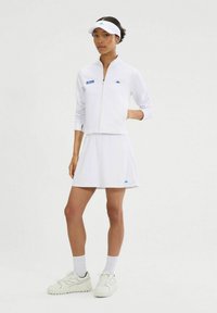Chaqueta deportiva blanca con cremallera, con un logo azul, combinada con una falda blanca y zapatillas blancas. La visera complementa el look deportivo.