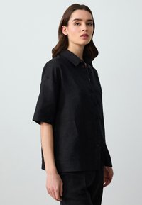 Camicia nera di lino a maniche corte con design a bottoni, colletto a punta e vestibilità rilassata. La texture del tessuto appare liscia e traspirante.