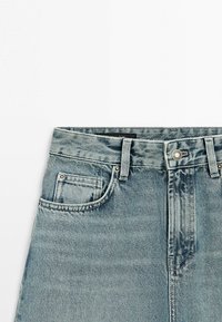 Lichtblauwe spijkerbroek taillegebied met voorste knoop, riemlussen en details van het voorvak op een witte achtergrond.