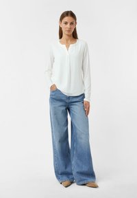 Die Frau steht in einer weißen Bluse mit langen Ärmeln und weiten blauen Jeans mit beigefarbenen spitzen Schuhen vor einem schlichten weißen Hintergrund.