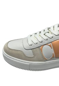 Sneaker classico con una tomaia in pelle bianca e dettagli in suede beige, lacci piatti di colore chiaro e un dettaglio del logo arancione sul lato.