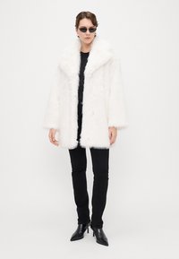 Cappotto in pelliccia sintetica bianco con ampio colletto, maniche oversize e una consistenza morbida. Indossato sopra una maglietta nera e abbinato a pantaloni neri e stivali.