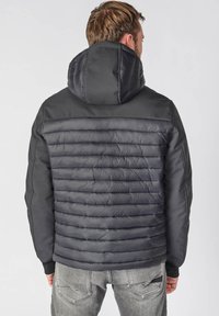 Zwarte pufferjack met capuchon, met gestikte delen op de rug en gladde schouders. Gemaakt van lichtgewicht, waterafstotendmateriaal.