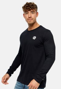Camisa negra de manga larga hecha de algodón suave, con un escote redondo y un pequeño logo blanco en el pecho. Diseño casual.