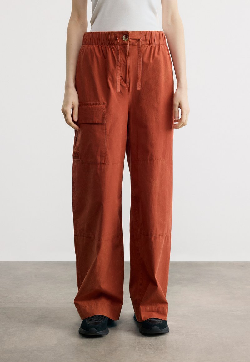 Personne portant un pantalon cargo ample de couleur rouille avec une poche latérale et une taille à cordon, associé à des baskets noires et un haut blanc.