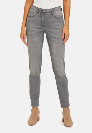 Frau trägt eine graue Slim-Fit-Jeans mit seitlichen und vorderen Taschen, kombiniert mit beigen Knöchelstiefeln vor einfarbigem Hintergrund.