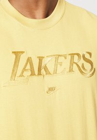 Camiseta amarilla de Nike con texto brillante en dorado "LAKERS" bordado en el pecho, usada por una persona con cuello visible y hombros parciales.