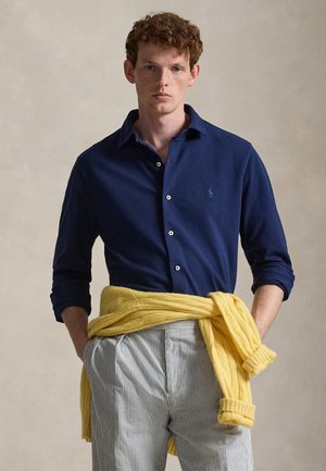 Junger Mann mit lockigem Haar, der ein dunkelblaues Hemd mit Knöpfen, gestreifte Hosen und einen gelben Pullover um die Taille gebunden trägt.