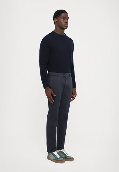 Homme debout portant un pull bleu marine ajusté, un pantalon gris foncé et des baskets vert-gris avec des lacets blancs sur un fond léger uni.