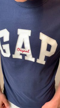 Marineblauwe katoenen T-shirt met grote witte letters die "GAP" spellen en rode cursieve "Original" eronder. Ronde hals en korte mouwen.