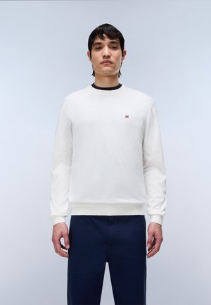 Mand iført hvid crewneck sweatshirt med lille norsk flaglogo og mørkeblå bukser, stående mod en ensfarvet lys baggrund.