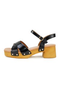 Zwarte leren sandaal met kruislussen en gouden studs, met een chunky houten hak en een gladde tan voetbed.