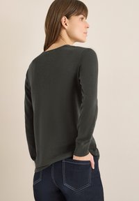 Ciemnozielona koszula z długim rękawem, okrągłym dekoltem i dopasowanymi mankietami, noszona z ciemnym dżinsem, ukazująca gładką fakturę materiału.