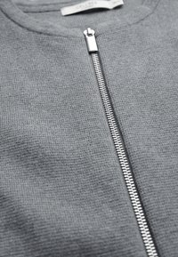 Grå zip-up sweater med rund halsudskæring, tekstureret stof og kontrasterende sølvlynlås. Stoffet ser blødt og strikket ud.