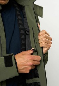 Olivroheline jakk, millel on lukuklipp ja must vooder, millel on korduv logo muster. Velcro mansetid ja lisahoidik.