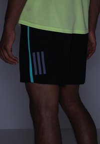 Pantalones cortos negros con una textura suave, que presentan una franja de color verde azulado a lo largo del costado y tres franjas verticales grises en la parte trasera.
