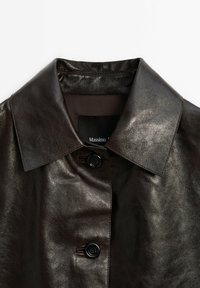 Veste en cuir marron foncé avec un large col et deux boutons noirs visibles, montrant une partie de l'étiquette intérieure avec le texte de la marque.
