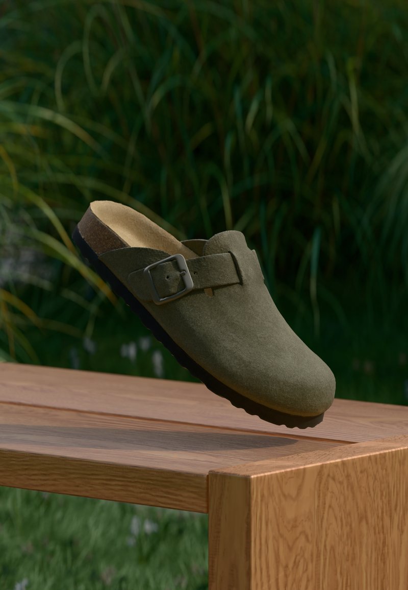 Grüne Wildleder-Mules mit einer abgerundeten Spitze, verstellbarem Metallverschluss und einem konturierten Korkfußbett. Strukturierte Sohle auf einer Holzoberfläche.