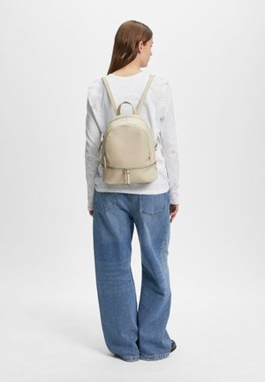 Frau trägt ein weißes Langarmshirt und blaue Jeans, sie trägt einen kleinen beigen Leder-Rucksack mit doppeltem Reißverschluss auf dem Rücken.