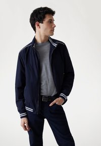 Marineblauwe ritsjack met witte strepenaccenten op de kraag en cuffs, gedragen over een grijs shirt, gecombineerd met donkere denimbroeken. Eenvoudig ontwerp.