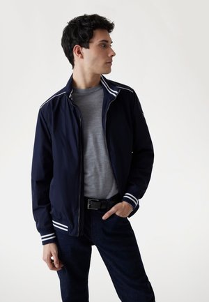 Giacca zip blu navy con dettagli a strisce bianche su colletto e polsini, indossata sopra una maglietta grigia, abbinata a pantaloni di jeans scuri. Design semplice.