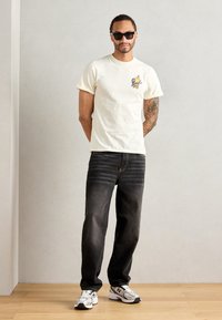 Lys cremefarvet bomulds t-shirt med grafisk print på brystet, parret med mørk denim jeans og grå sportsko med hvide detaljer.