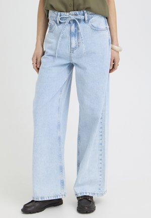 Žena nosí svetlomodré široké džínsy s vysokým pásom a viazaným opaskom, olivovú košeľu, čierne topánky a biely náramok na pravom zápästí.