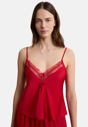Kobieta w czerwonej satynowej koszulce na ramiączkach z koronkowymi wykończeniami i kokardkami, widoczna od połowy tułowia w górę na jednolitym tle.