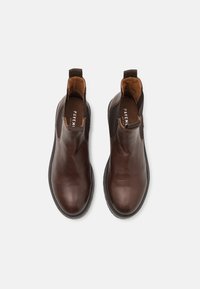 Botas Chelsea de cuero marrón con paneles laterales elásticos, textura suave, punta redonda y suela de goma oscura en contraste. Nombre de la marca visible en el interior.