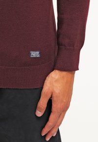 Bordeauxfarbener Strickpullover mit gerippten Bündchen und Saum, versehen mit einem kleinen blauen Etikett mit weißem Text auf der unteren linken Seite in der Nähe der Naht.