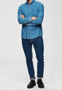 Muž stojící se založenýma rukama, oblečený v modré denimové košili, tmavě modrých ohrnutých džínách a černých kožených botách na jednotném pozadí.